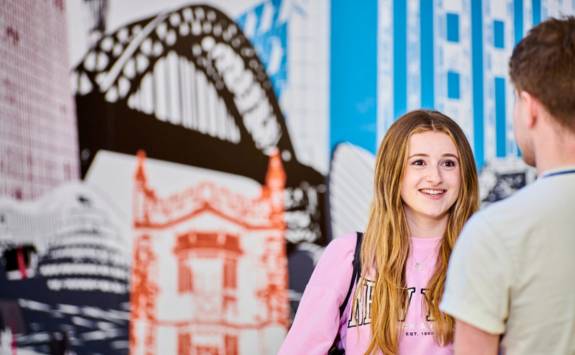 Two NUBS undergraduates standing in the Business School foyer. Behind them is a mural featuring the Tyne Bridge.